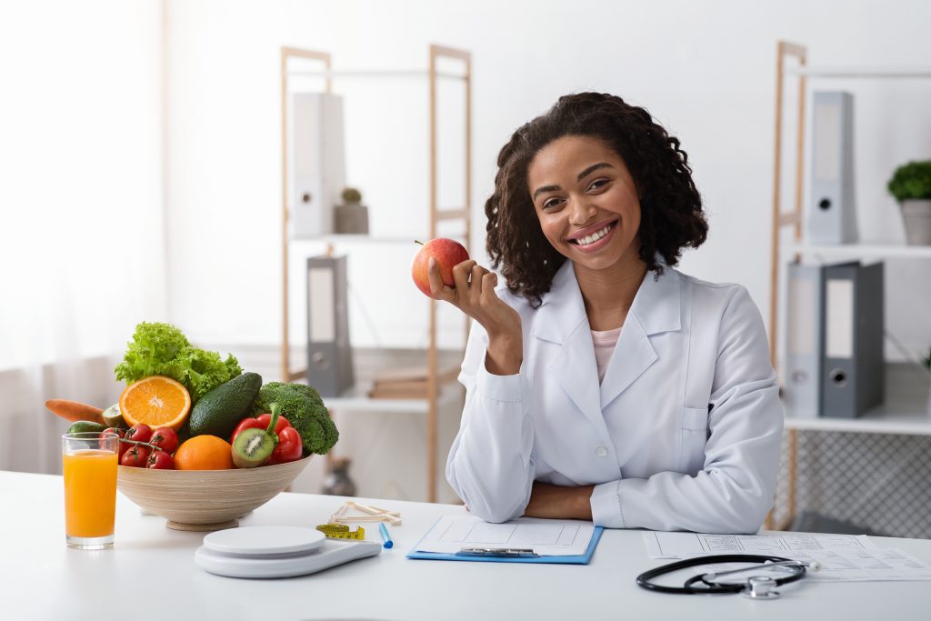 Jeune femme souriante en tenue de personnel médical, assise sur un bureau, symbolisant le professionnalisme et le bien-être dans le secteur de la santé, présenté sur madagasy.com.