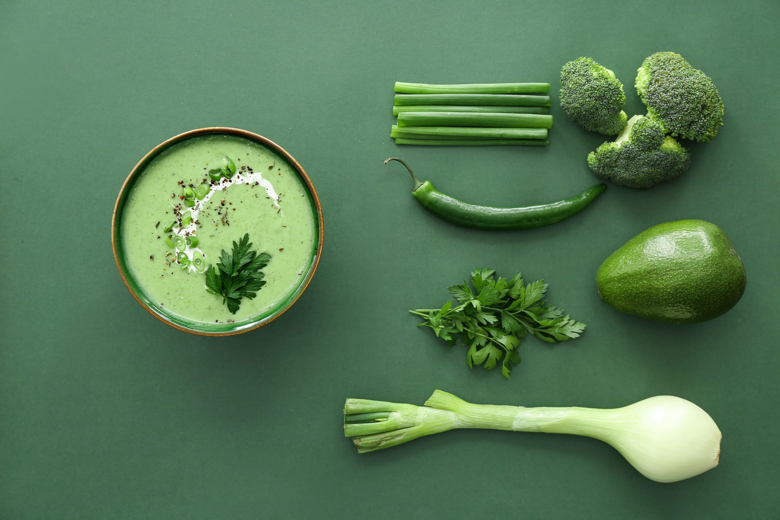 Un assortiment de légumes verts et un bol de soupe verte crémeuse, garni de persil, de crème et de poivre noir. Les légumes comprennent du brocoli, des oignons verts, un piment vert, un avocat, du persil et un poireau. Une composition mettant en avant des ingrédients frais et sains, présentée sur la plateforme Restauration de Madagasy.com