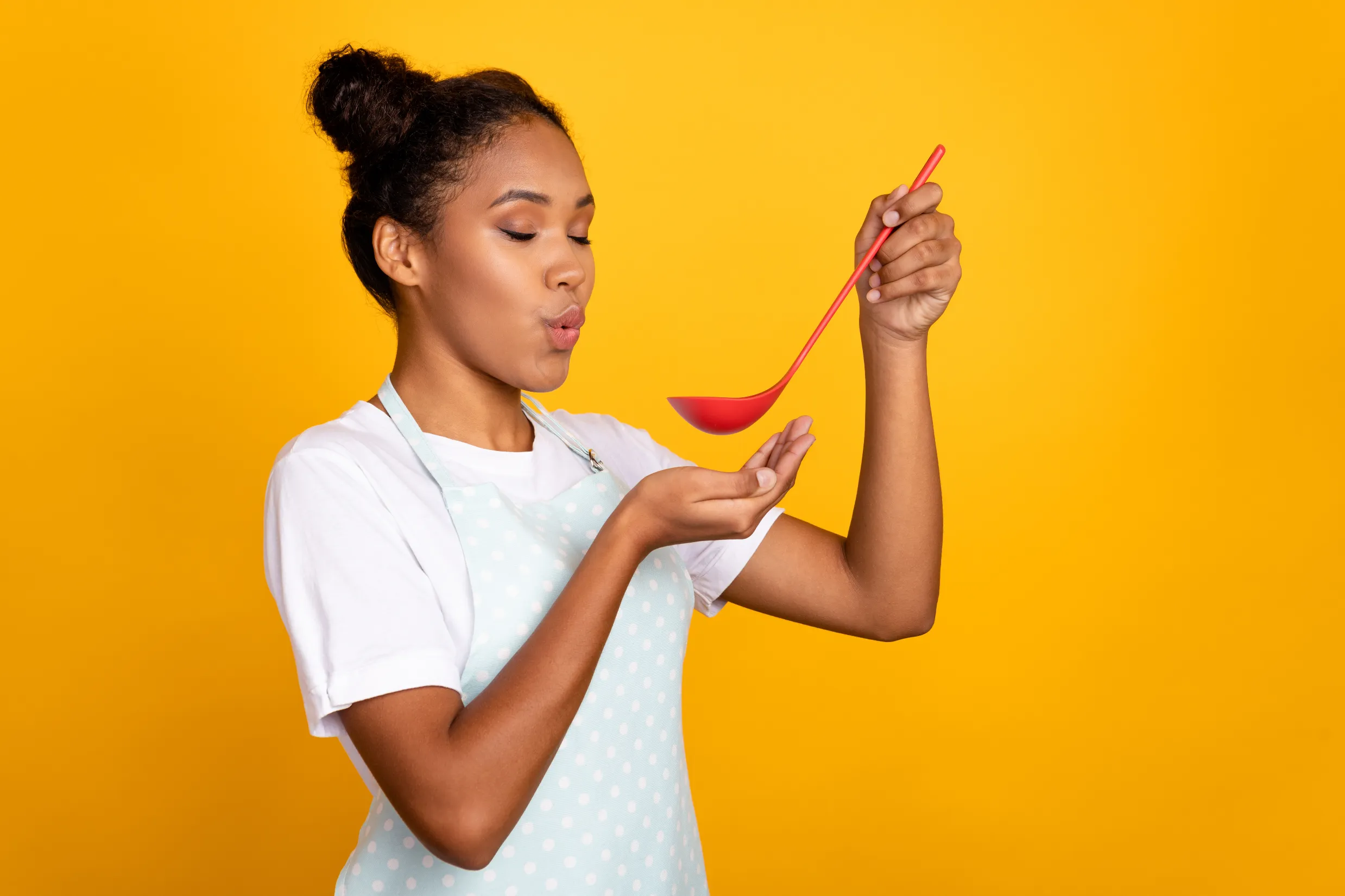 Une femme en tablier à pois blancs et t-shirt blanc, tenant une grande louche rouge au-dessus de sa main ouverte, sur un fond jaune vif. L'image évoque l'idée de présentation, de mise en valeur d'un produit ou d'une offre commerciale sur la plateforme Commerce de Madagasy.com, avec une posture engageante et dynamique.